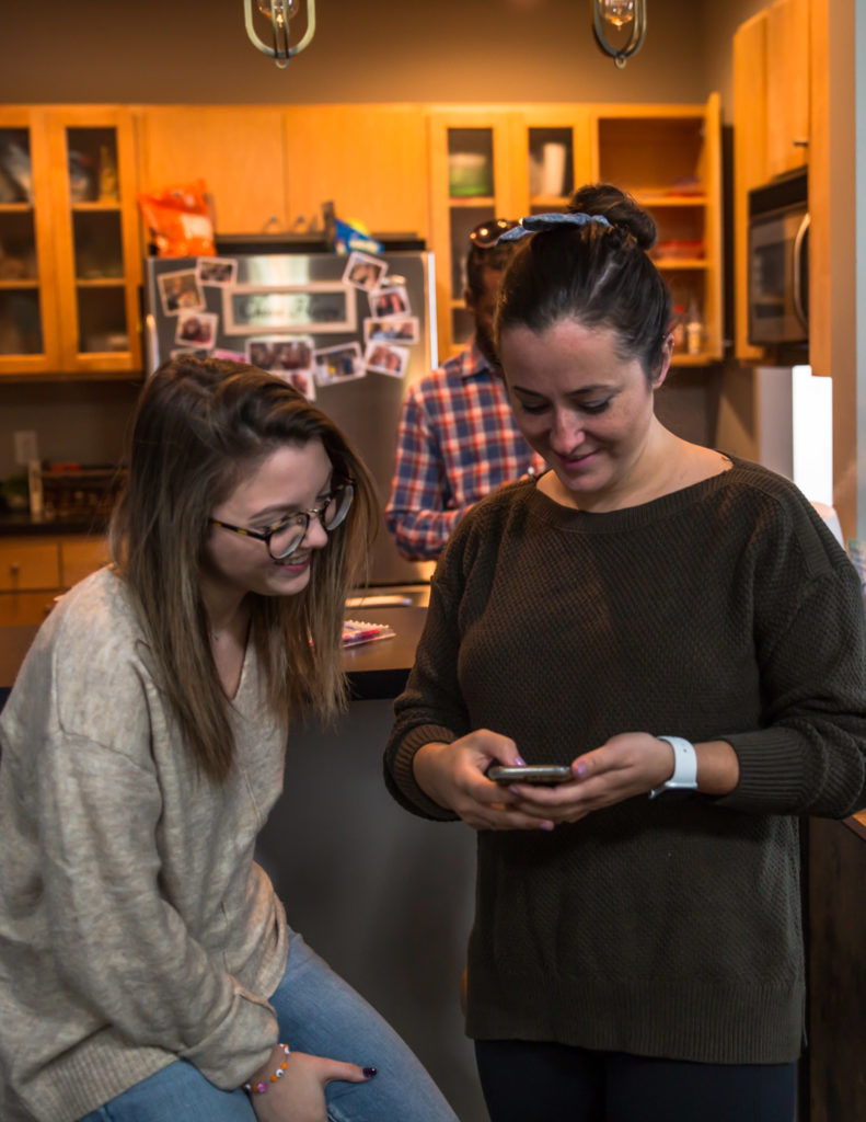 Two women in a kitchen looking at a cell phone.