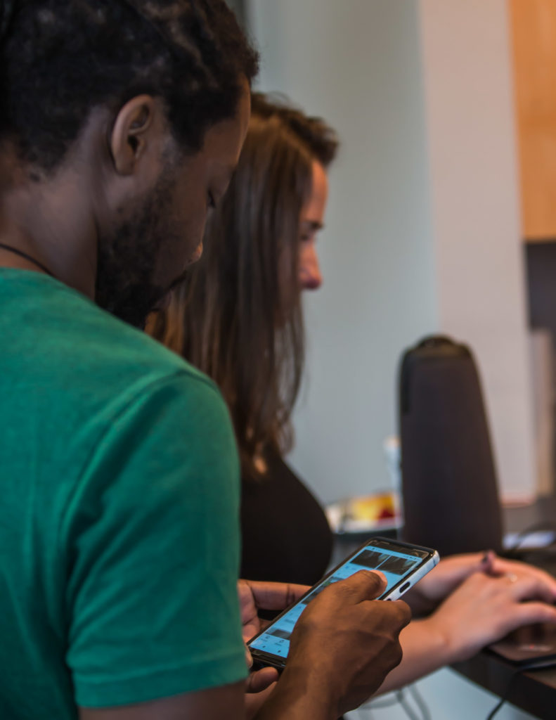 A man focused on his phone in the foreground, a woman on her laptop in the background.