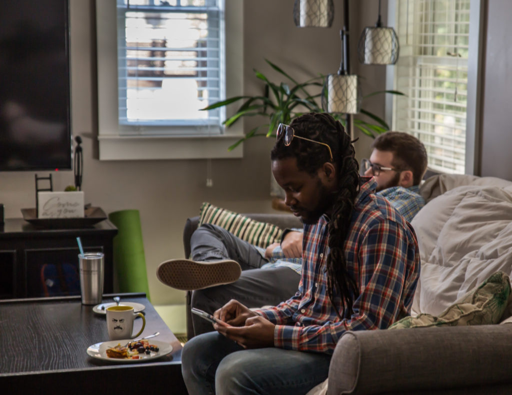 A man sitting comfortably on a couch at home while he looks at his phone next to another man.