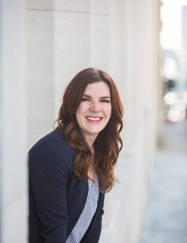 Headshot of Anna-Vija McClain who wear a striped blouse and navy blazer. She is a pale red-headed woman with long hair.