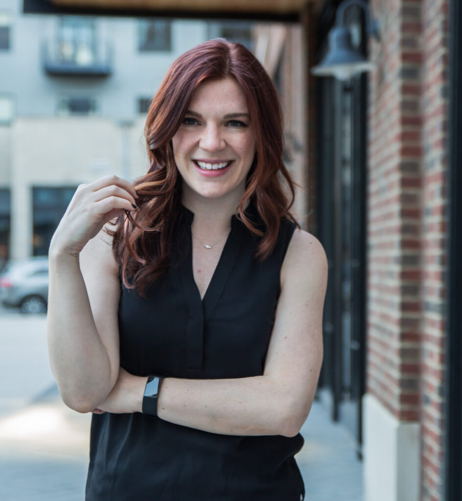 Anna-Vija McClain is a smiling woman with red hair standing confidently on an urban street, dressed in a stylish black sleeveless top.