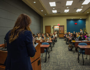 Anna-Vija McClain, a woman with long red hair, acting as a speaker. She is facing an attentive audience during a seminar in a conference room.