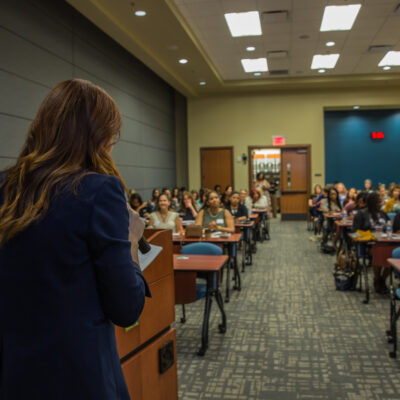 Anna-Vija McClain, a woman with long red hair, acting as a speaker. She is facing an attentive audience during a seminar in a conference room.