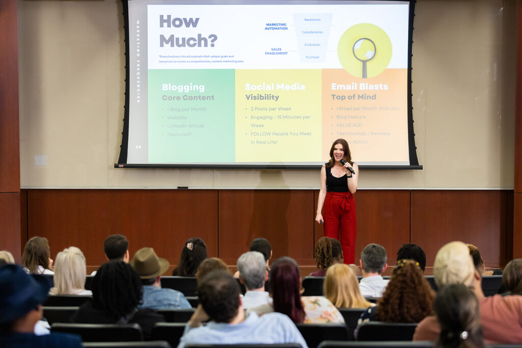 Anna-Vija McClain presents to an attentive audience at a seminar, with a presentation slide highlighting strategies for blogging, social media visibility, and email blasts.