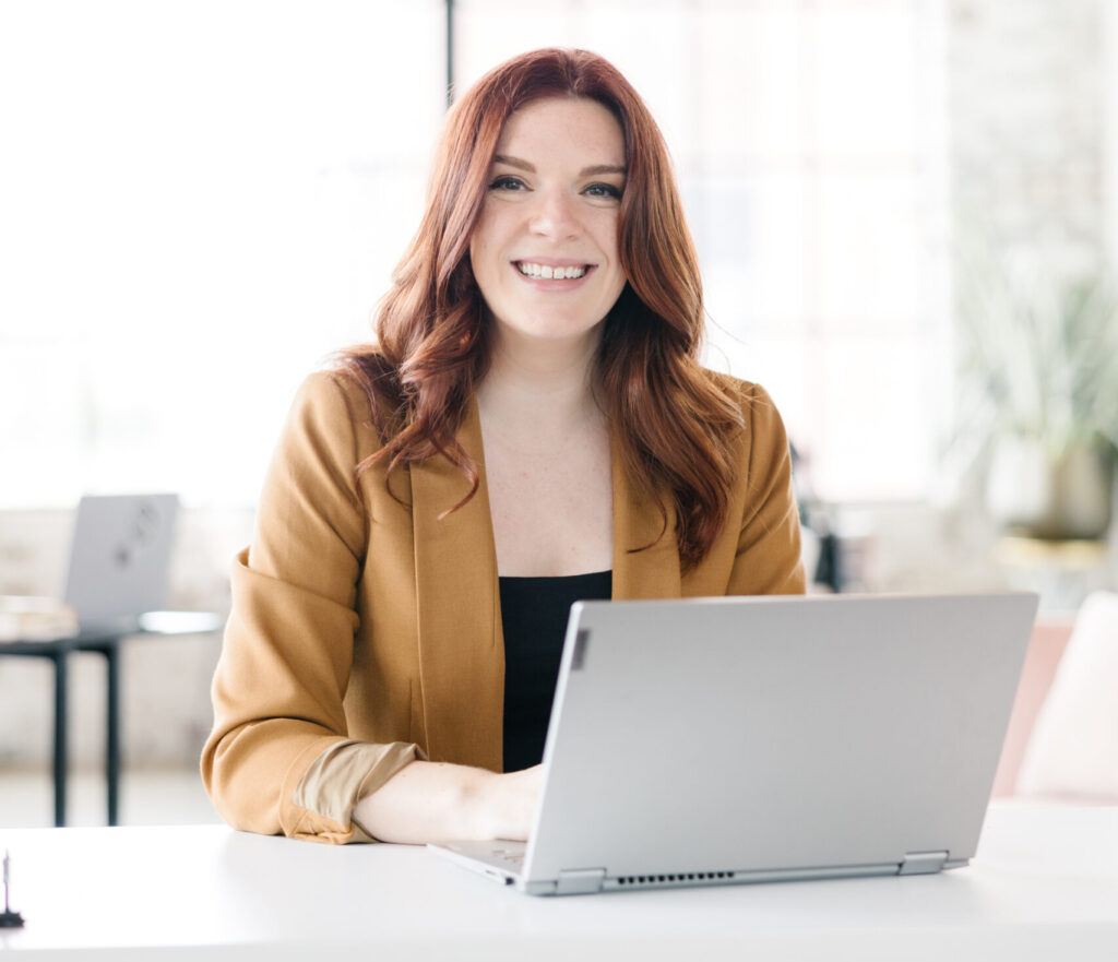 Anna-Vija McClain, a smiling woman with red hair wearing a mustard-colored blazer sits at a desk behind her laptop in a bright, modern office.